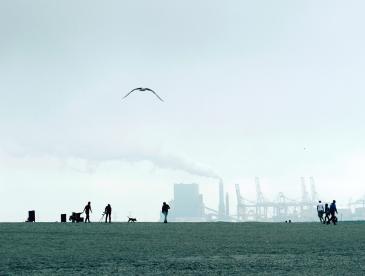 Distant view of people in a field, with a factory in the background releasing smoke.